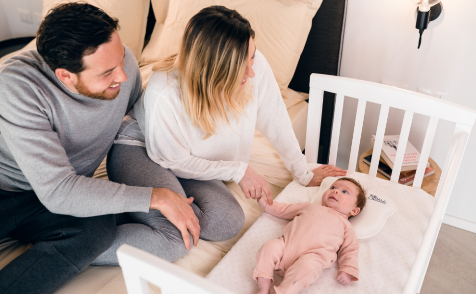 Parents watch baby girl in the crib next to the bed with a white plagiocephaly baby pillow