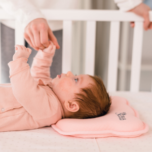 Newborn baby lying in crib with plagiocephaly pillow in pink takes her mum's hand