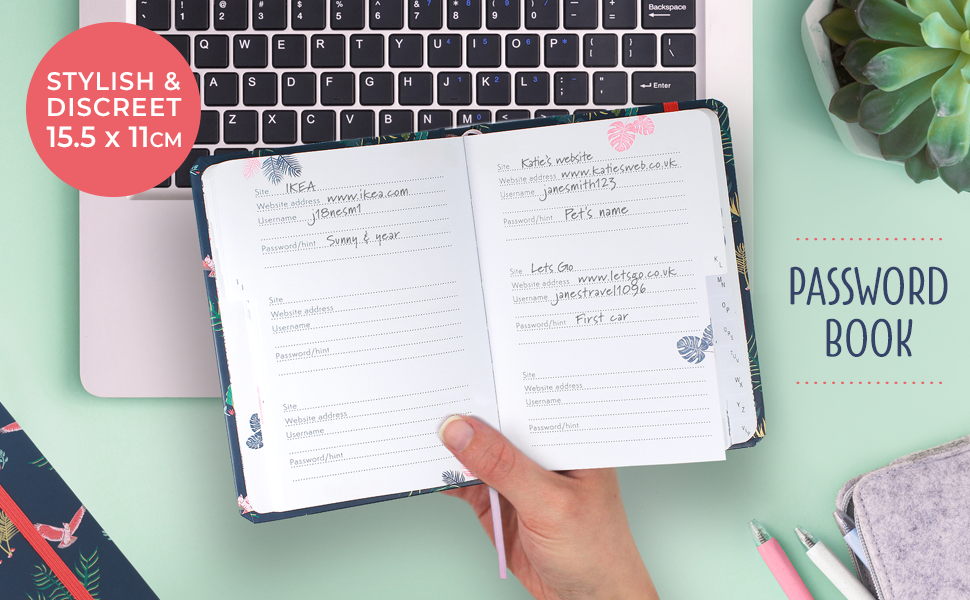 A person holding an open password book in front of their laptop keyboard, pens and an indoor plant