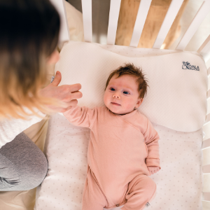 Mum takes her daughter's hand; infant is lying in the crib with a Maxi Plagiocephaly baby pillow 